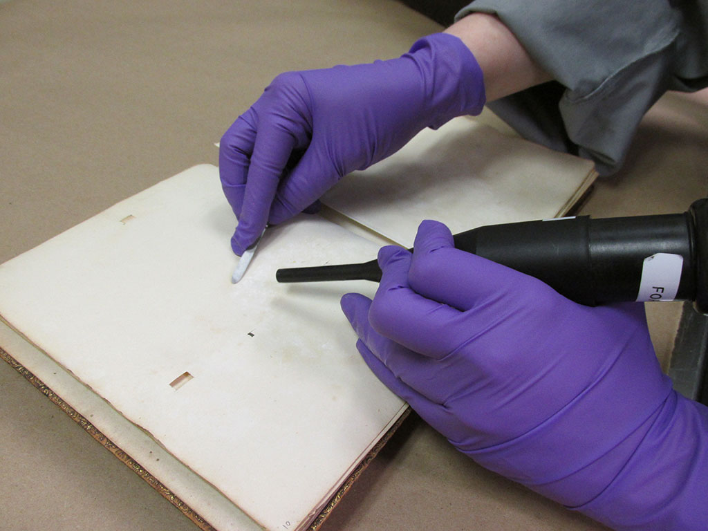 Removing the white powder using a HEPA-filtered vacuum. The book is not considered fully decontaminated, even after vacuuming and surface cleaning, so the conservator and photographer needed to wear PPE throughout the project.